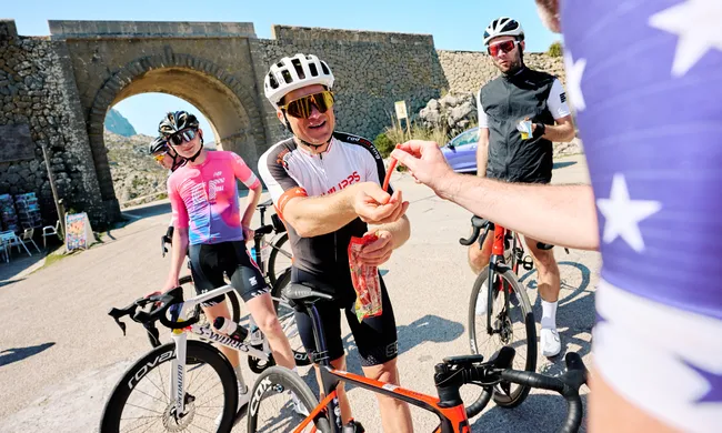 Radfahrer machen eine Pause am Sa Calobra Krawattenknoten. Ein Mann reicht einem anderen Süßigkeiten, während andere daneben stehen.