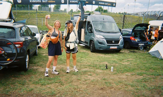 Zwei junge Frauen jubeln auf einem grünen Festivalgelände mit Autos, Camper und Zelt im Hintergrund.