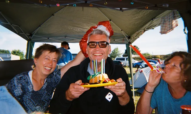 Drei lächelnde Personen feiern Geburtstag im Freien unter einem Zelt. Ein Mann in der Mitte hält einen Kuchen, daneben eine Frau mit Partytröte.