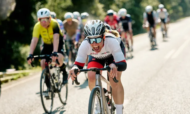 Yvonne Margraf, eine Radfahrerin in weiß-rotem Trikot lacht vor Freude beim Rennen auf sonniger Straße, andere Radfahrer unscharf im Hintergrund.