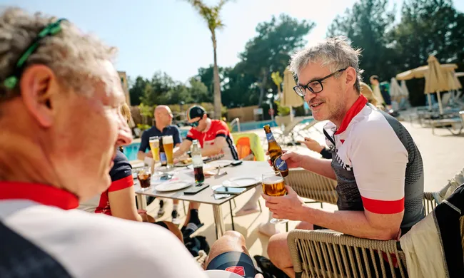 Radfahrer machen auf einer sonnigen Terrasse mit Getränken Pause. Ein Mann in Radkleidung hält ein Bier und lächelt.