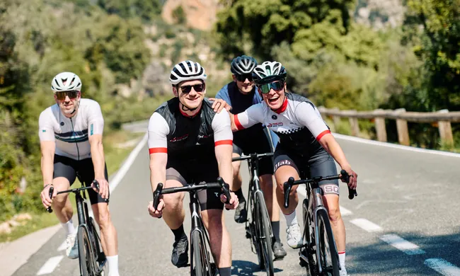 Vier glückliche Radfahrer in Sportkleidung fahren auf einer sonnigen Straße mit grünen Hügeln im Hintergrund.