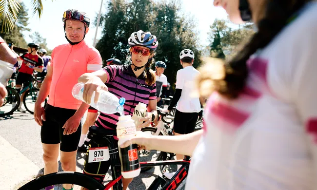 Radfahrer füllen bei strahlendem Sonnenschein Wasserflaschen nach. Eine Frau in gestreiftem Trikot gießt Wasser um, ein Mann in Korallentrikot lächelt.