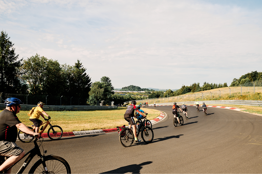Cyclists ride on a curving track surrounded by trees and grassy fields under a partly cloudy sky.