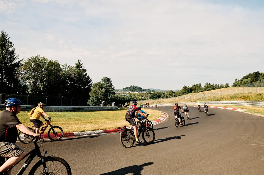 Cyclists ride on a curving track surrounded by trees and grassy fields under a partly cloudy sky.