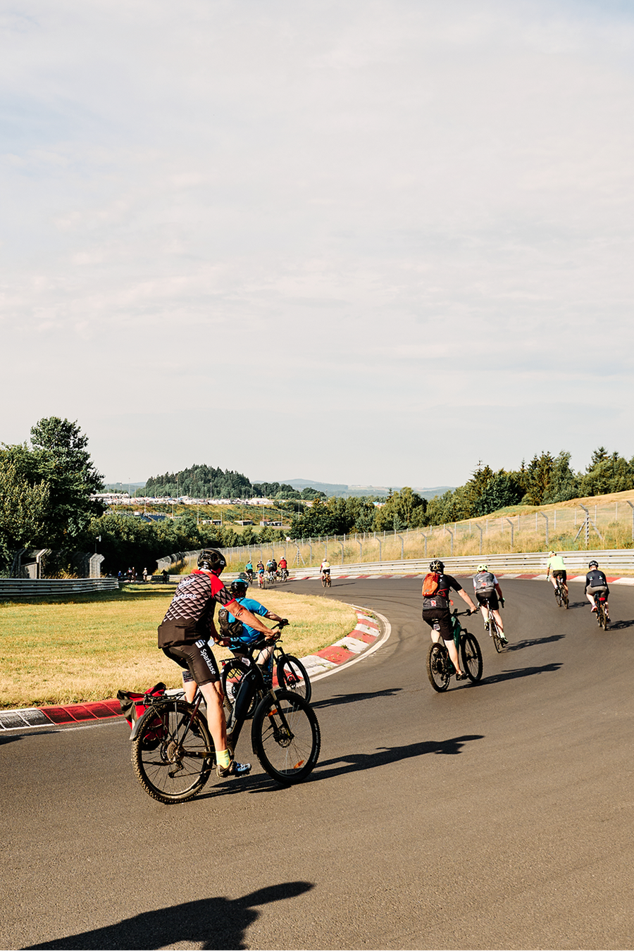 Cyclists ride on a curving track surrounded by trees and grassy fields under a partly cloudy sky.