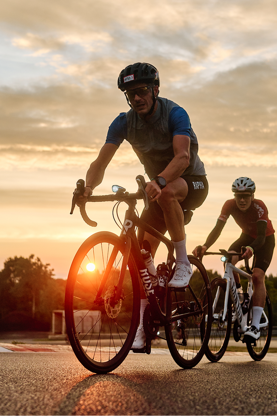 Cyclists riding on a road at sunset, with a glowing sky and trees in the background.
