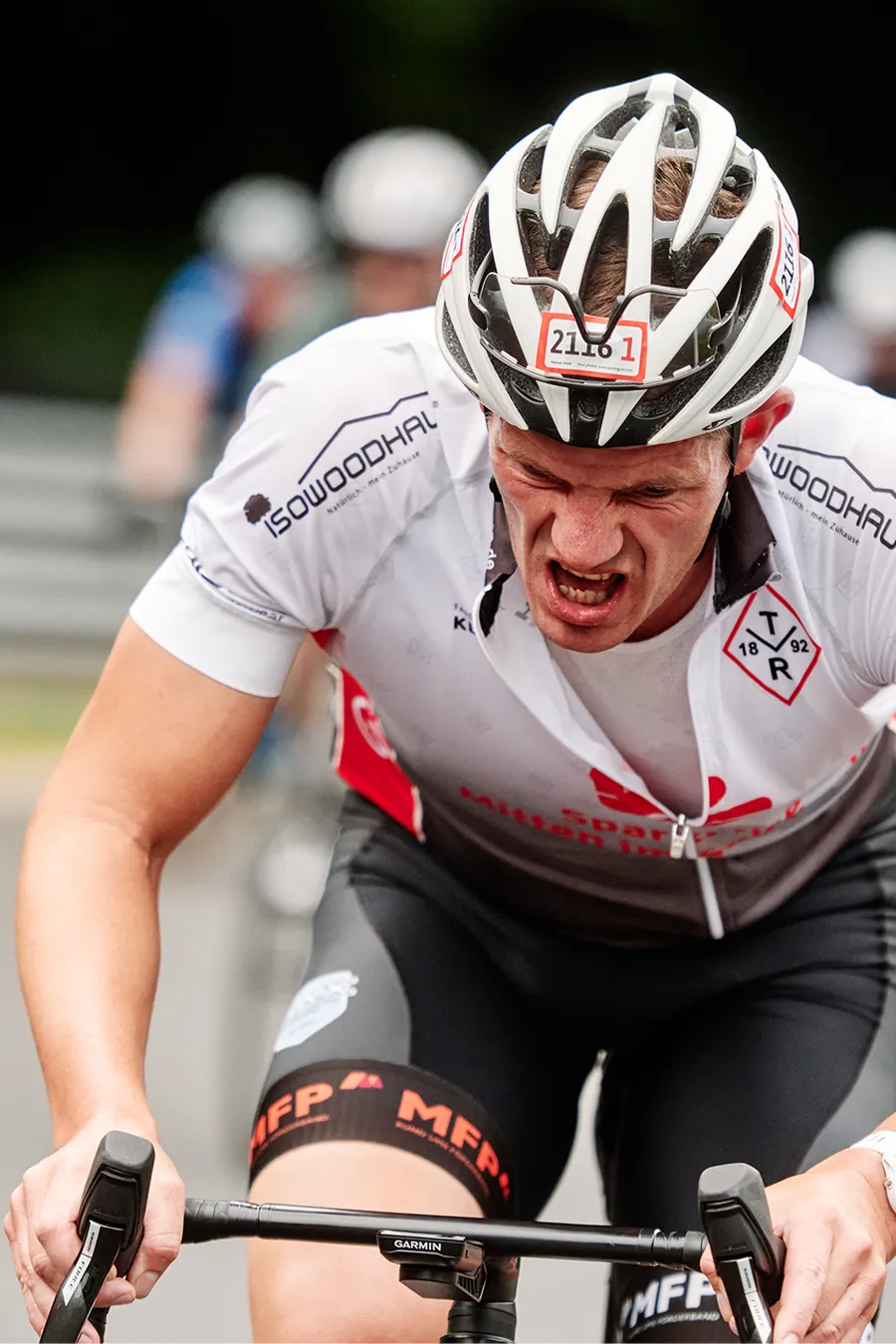 Male cyclist grimacing fiercely in a white helmet with number 2116, wearing a white and black cycling kit, riding hard on a road.