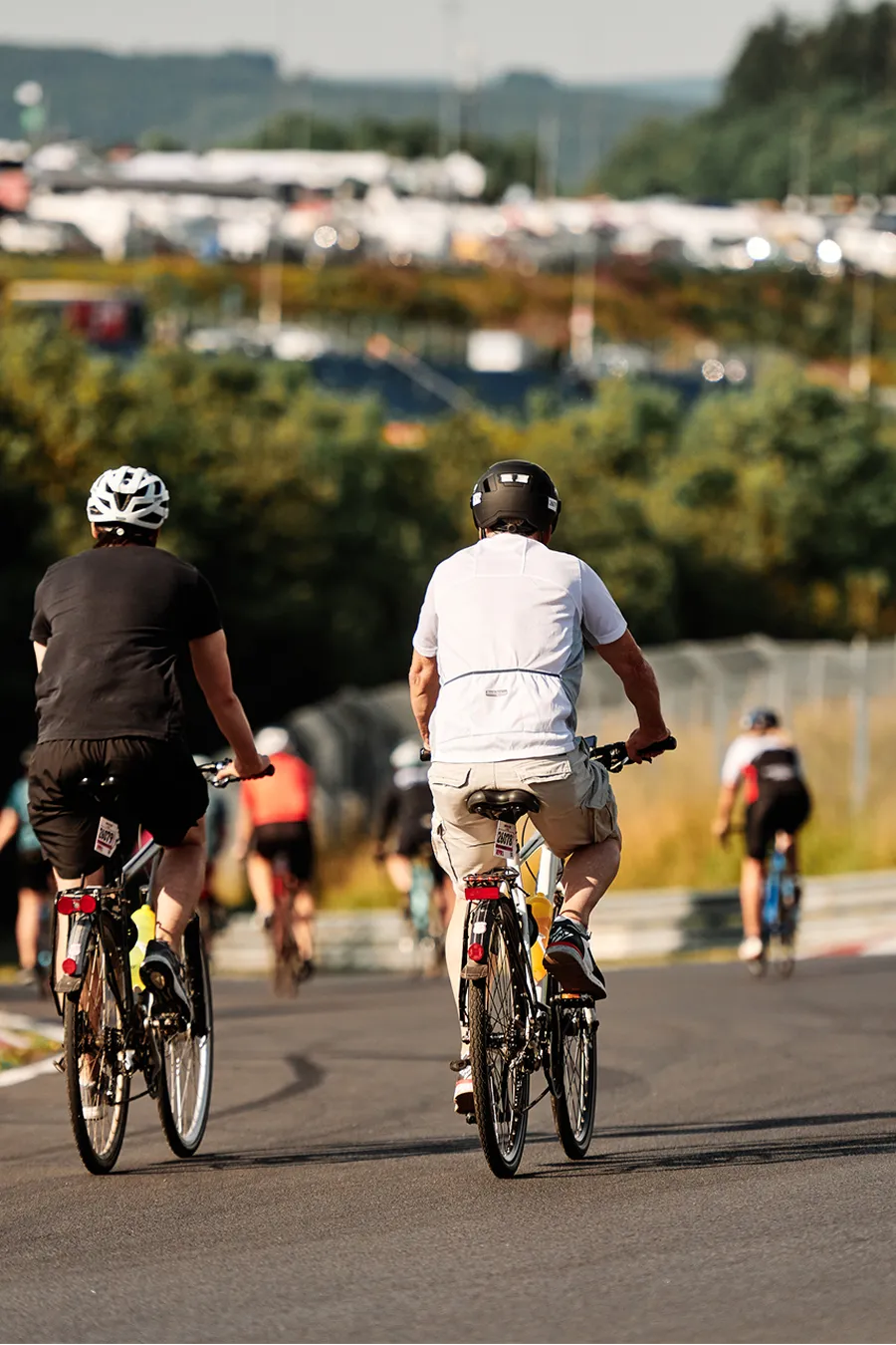 A group of cyclists ride on a paved circuit, viewed from behind. Two men in black and white shirts lead, with hills and a large camp site in the background.
