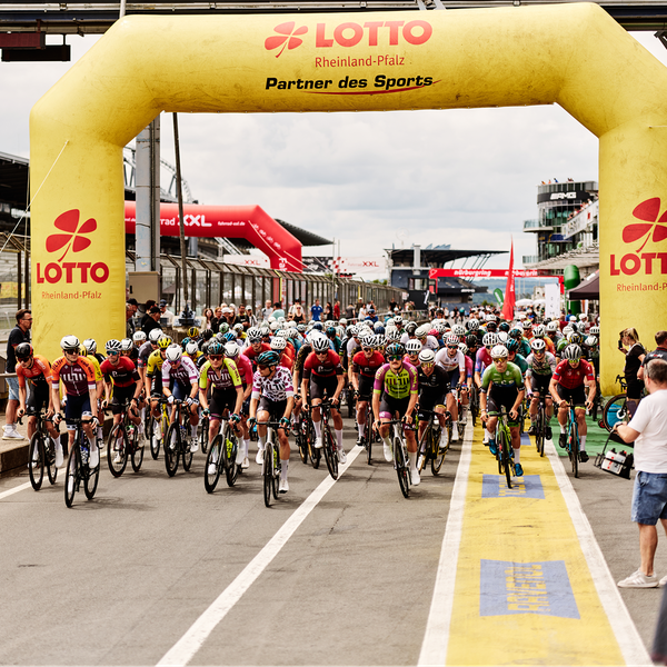 Cyclists race under a yellow inflatable archway at the start line of a cycling event, surrounded by spectators and a race track environment.