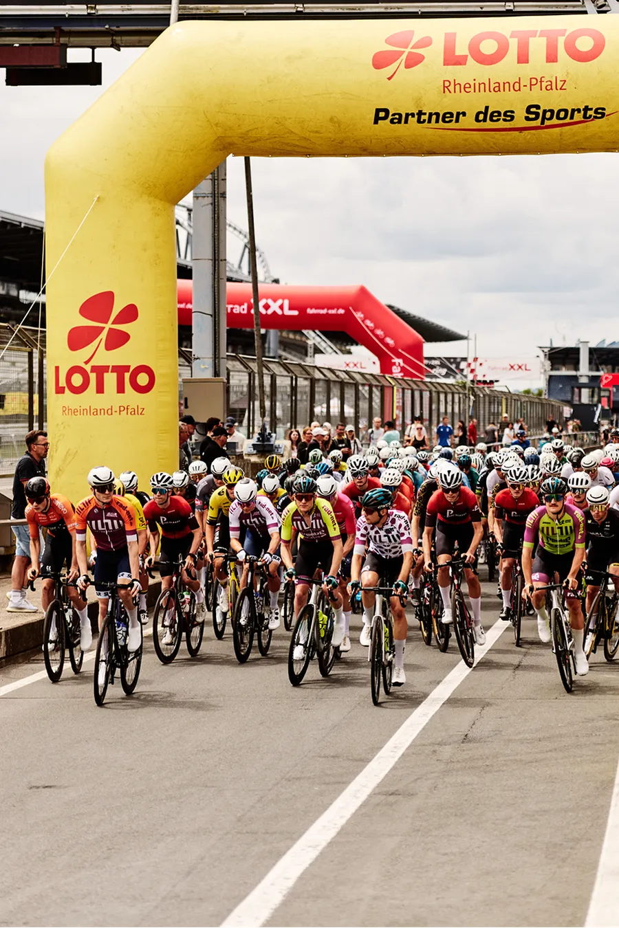 Cyclists race under a yellow inflatable archway at the start line of a cycling event, surrounded by spectators and a race track environment.
