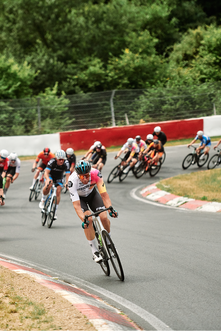 Cyclists race around a curved track, surrounded by green trees and a red and white barrier, with one rider leading the pack.