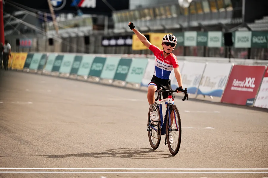 A triumphant young cyclist in a red, white, and blue uniform raises a fist, celebrating victory on a race track.