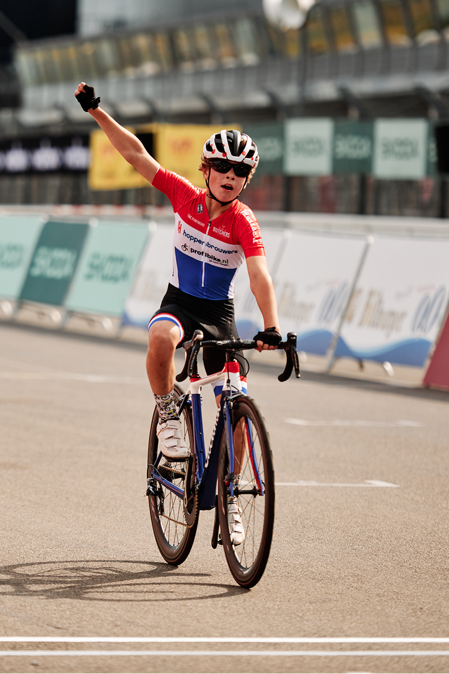 A triumphant young cyclist in a red, white, and blue uniform raises a fist, celebrating victory on a race track.