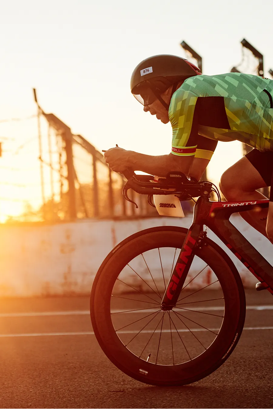 Cyclist in a race riding a bike at sunset, wearing a green jersey and helmet, passing a barrier with sun flaring in the background.