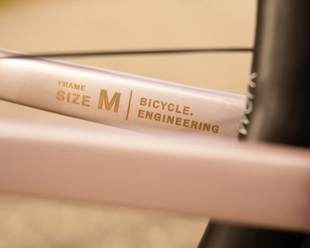 Nahaufnahme eines roségoldenen Fahrradrahmens mit goldenem Text "FRAME SIZE M" und "BICYCLE. ENGINEERING".