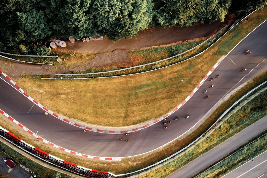Nordschleife Drohnenfoto Aerial view of cyclists on a winding road surrounded by greenery, with a red and white curb lining the curve.