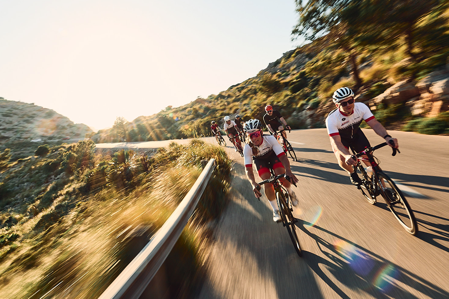 TOUR Trainingscamp A group of cyclists speeds along a winding mountain road with motion blur. Bright sunlight flares from the horizon, casting long shadows.