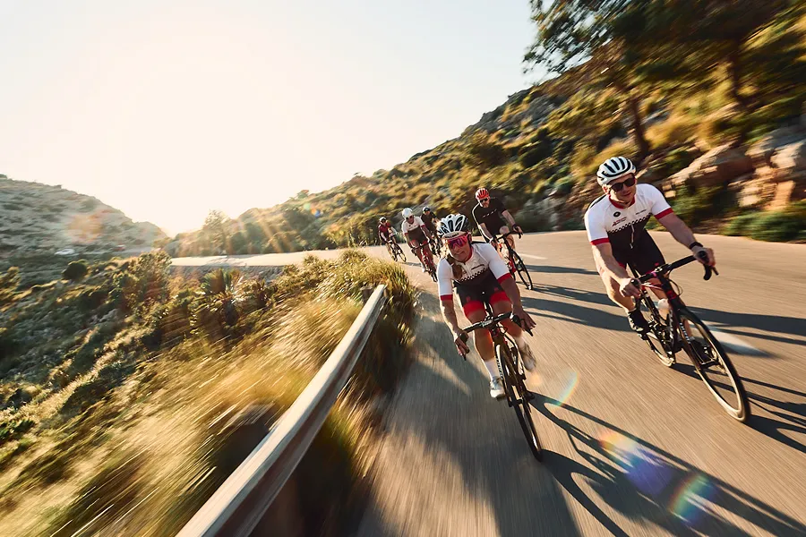 A group of cyclists speeds along a winding mountain road with motion blur. Bright sunlight flares from the horizon, casting long shadows.