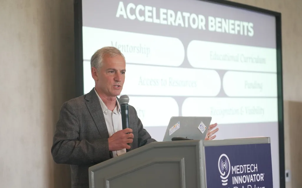 A speaker at a MedTech Innovator Radar Forum podium presenting a slide on accelerator benefits