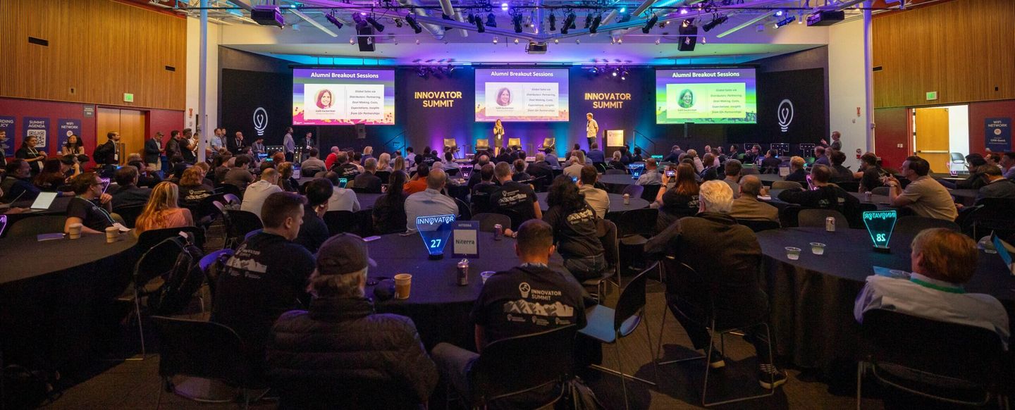 Main hall of the Innovator Summit 2024 at the Computer History Museum, with attendees at round tables facing the stage screens