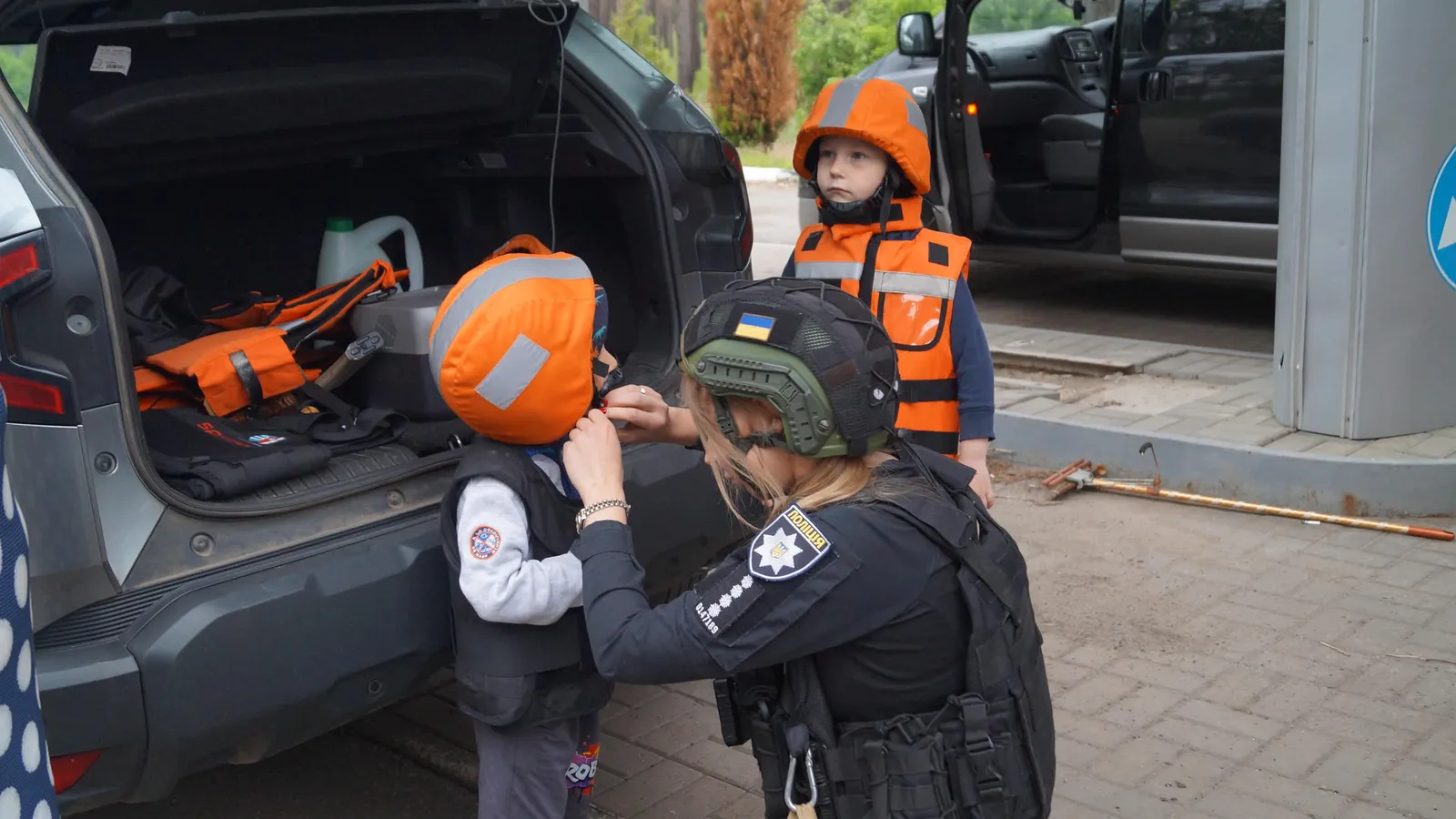 En politibetjent spænder en beskyttelsesvest på et barn med orange hjelm ved et åbent bagagerum