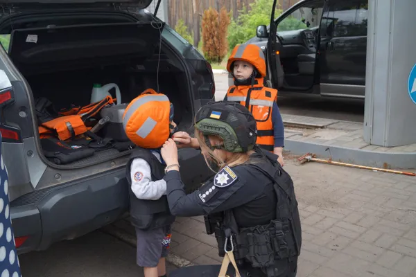 En politibetjent spænder en beskyttelsesvest på et barn med orange hjelm ved et åbent bagagerum