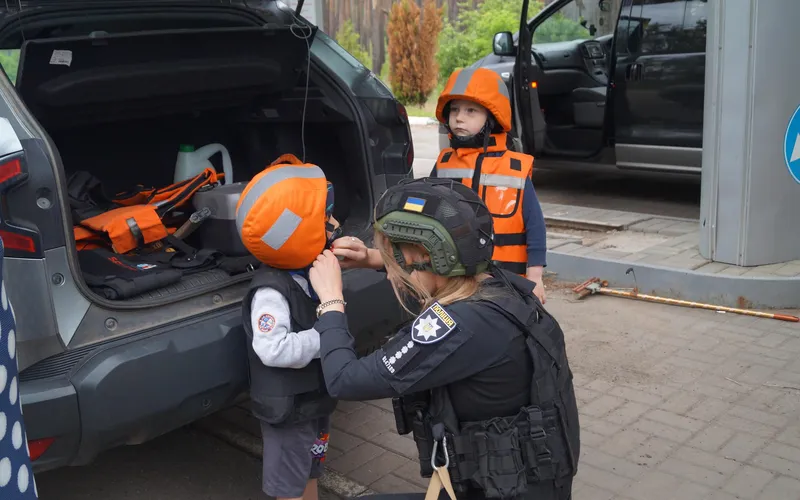 En politibetjent spænder en beskyttelsesvest på et barn med orange hjelm ved et åbent bagagerum