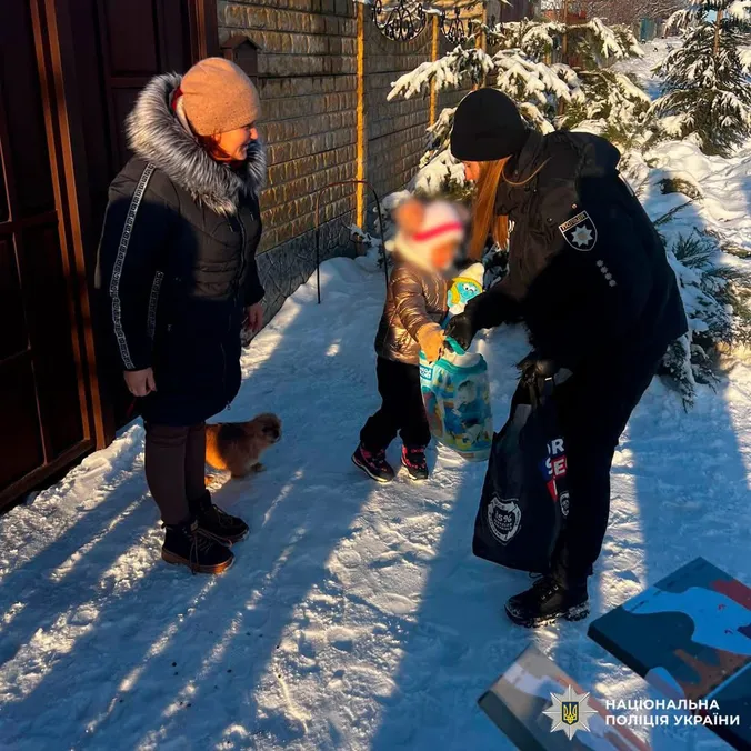 Politibetjent rækker en pose og en gave til et lille barn i en snedækket have, mens moren ser til.