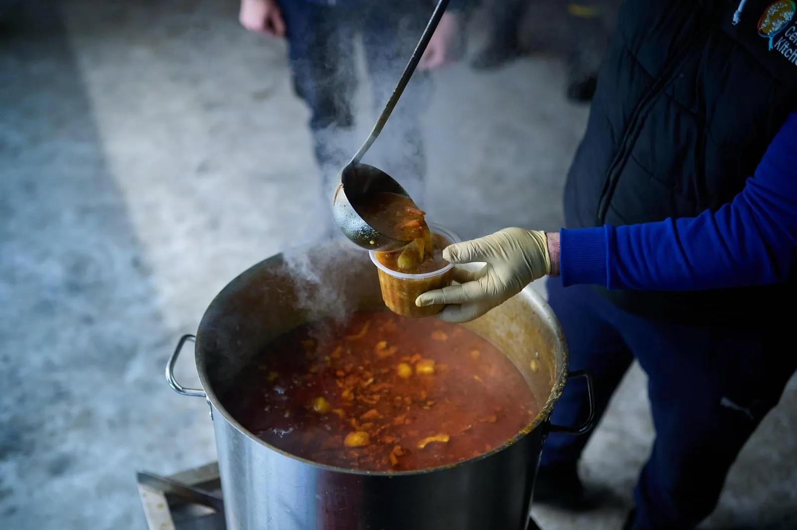 Behandsket hånd øser dampende suppe op i et bæger til uddeling