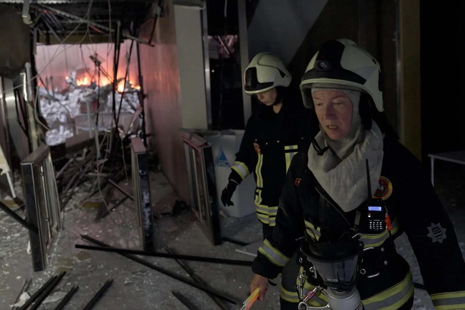 To brandfolk med hjelme går gennem en udbrændt bygning, hvor flammerne stadig brænder i baggrunden.