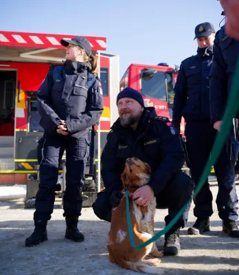 Uniformeret mand sidder på hug med en hund i snor foran en rød brandbil i sneen.