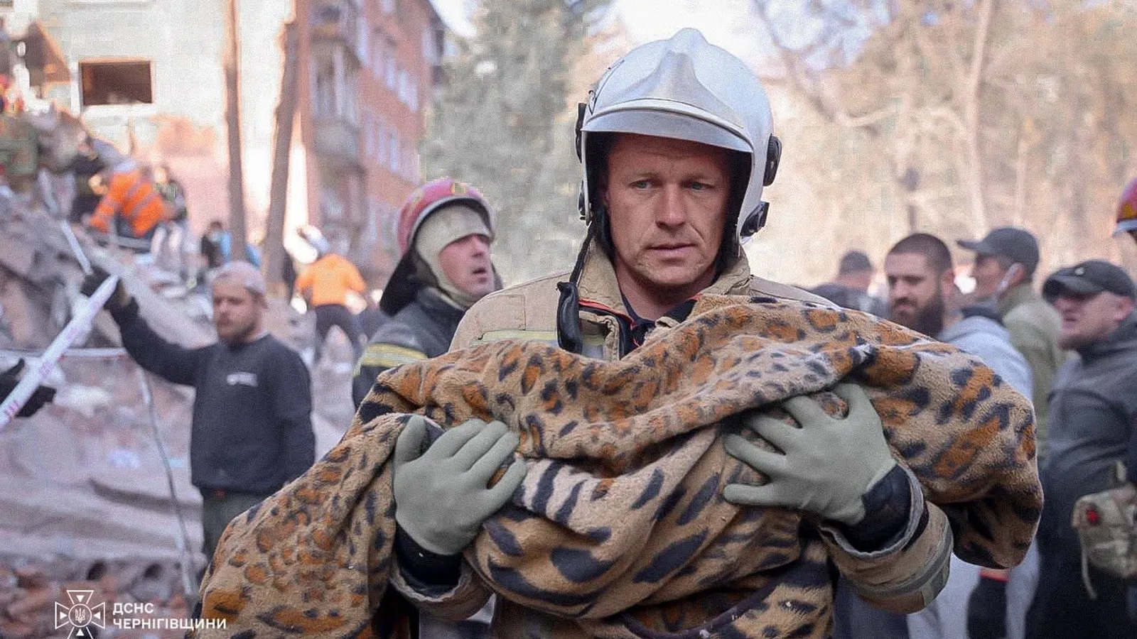 Ukrainsk redningsmand bærer et indsvøbt tæppe væk fra den sønderbombede boligblok i Pryluky, mens redningsfolk arbejder i baggrunden