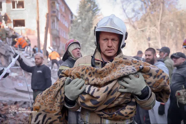 Ukrainsk redningsmand bærer et indsvøbt tæppe væk fra den sønderbombede boligblok i Pryluky, mens redningsfolk arbejder i baggrunden
