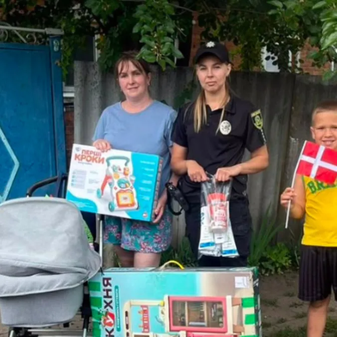 Kvinde, ukrainsk politibetjent og dreng med lille dansk flag står ved en barnevogn og udleveret legetøj.