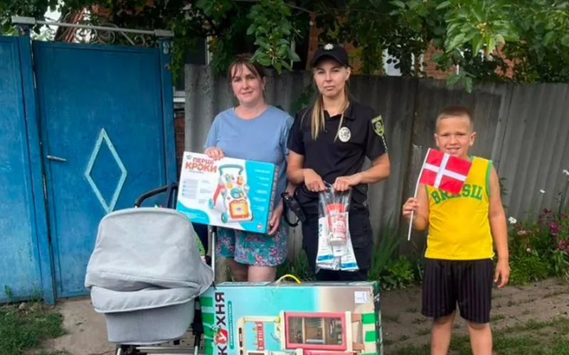 Kvinde, ukrainsk politibetjent og dreng med lille dansk flag står ved en barnevogn og udleveret legetøj.