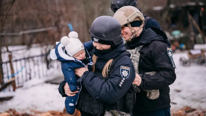 En politibetjent med hjelm bærer en baby i hvid hue, mens en anden betjent står ved siden af i sneen