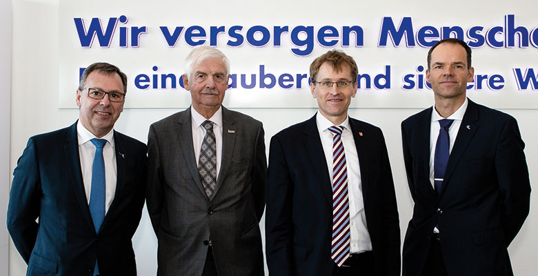 Vier Männer im Anzug vor dem Schriftzug Wir versorgen Menschen