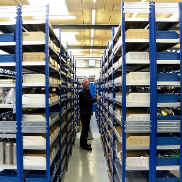 Person browsing items in a warehouse aisle, surrounded by tall blue metal shelves filled with boxes and various materials.