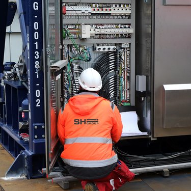 A worker in an orange safety jacket and white helmet kneels in front of an open electrical panel, inspecting cables and components.