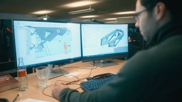 Person working at a desk with dual monitors displaying 3D design software, a keyboard, a glass, and a bottle nearby.