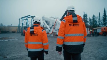 Two people in orange safety jackets and safety hats walk towards an a-frame on a work site.