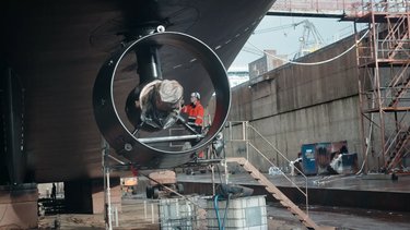 Workers inspecting equipment inside a large cylindrical structure on a ship in a shipyard, surrounded by scaffolding and tools.