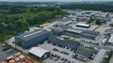 Aerial view of an industrial area with large buildings, numerous parked vehicles, and surrounding green areas.
