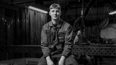 A man in work attire sits in an industrial workshop, holding a tool, with metal equipment and a soft light illuminating the background.