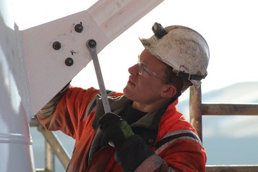 Construction worker in an orange jacket and helmet using a wrench to tighten bolts on a metal structure, with safety glasses on.