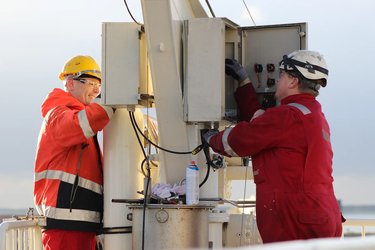 Two workers in protective clothing and helmets perform maintenance on an electrical box outdoors.