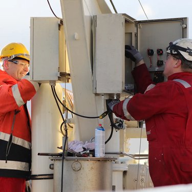 Two workers in protective clothing and helmets perform maintenance on an electrical box outdoors.