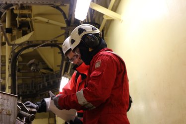 Two workers in protective gear and helmets examining documents in an industrial setting with overhead machinery and yellow walls.