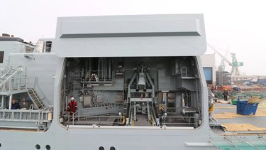 A large, detailed view of a ship's machinery compartment, with an opened hangar port, visible equipment and a person in a red uniform inside. Industrial background.
